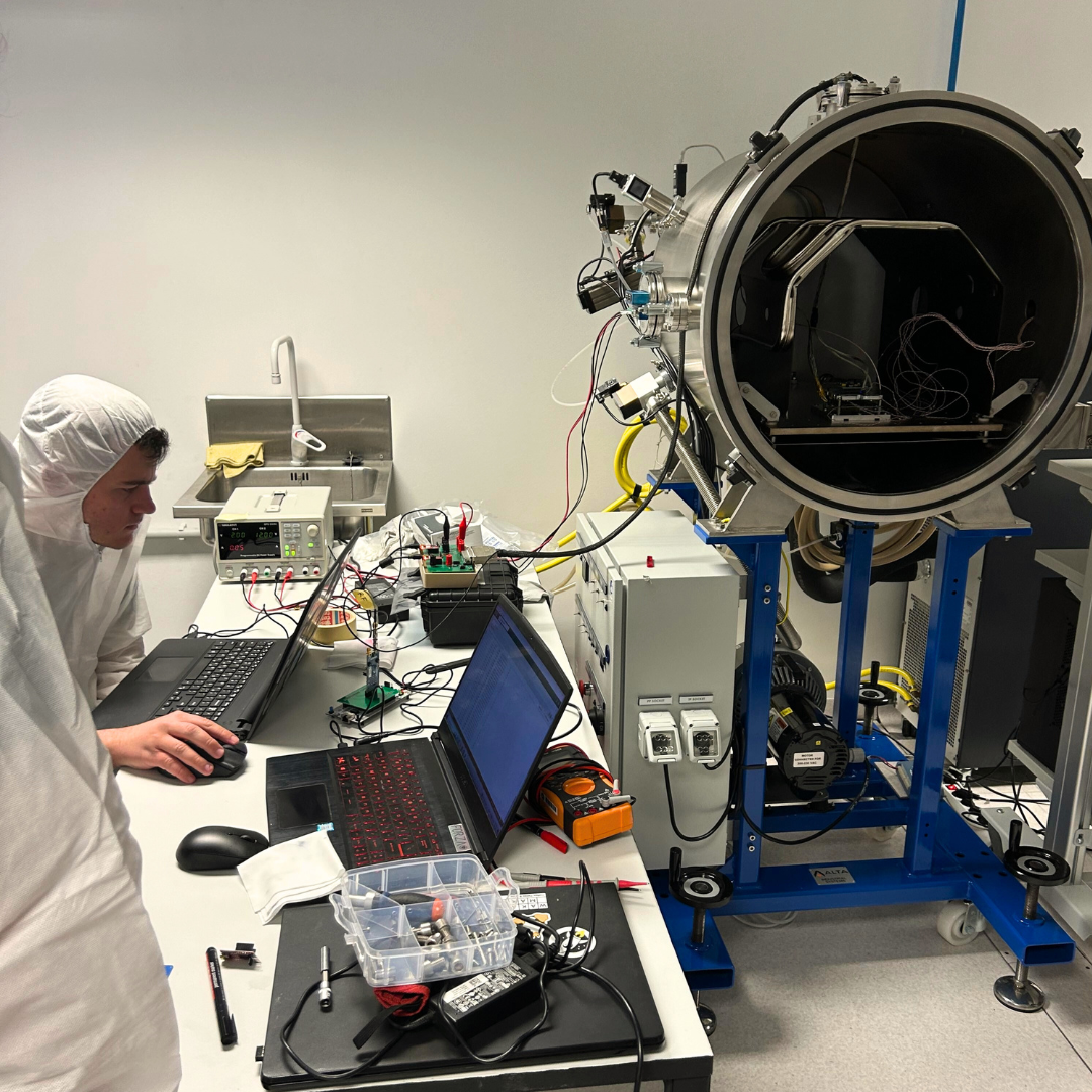 The PeakSat team working in a cleanroom. The PCB stack is placed inside an open Thermal Vacuum Chamber, while an engineer outside is configuring the electrical support equipment on a laptop.